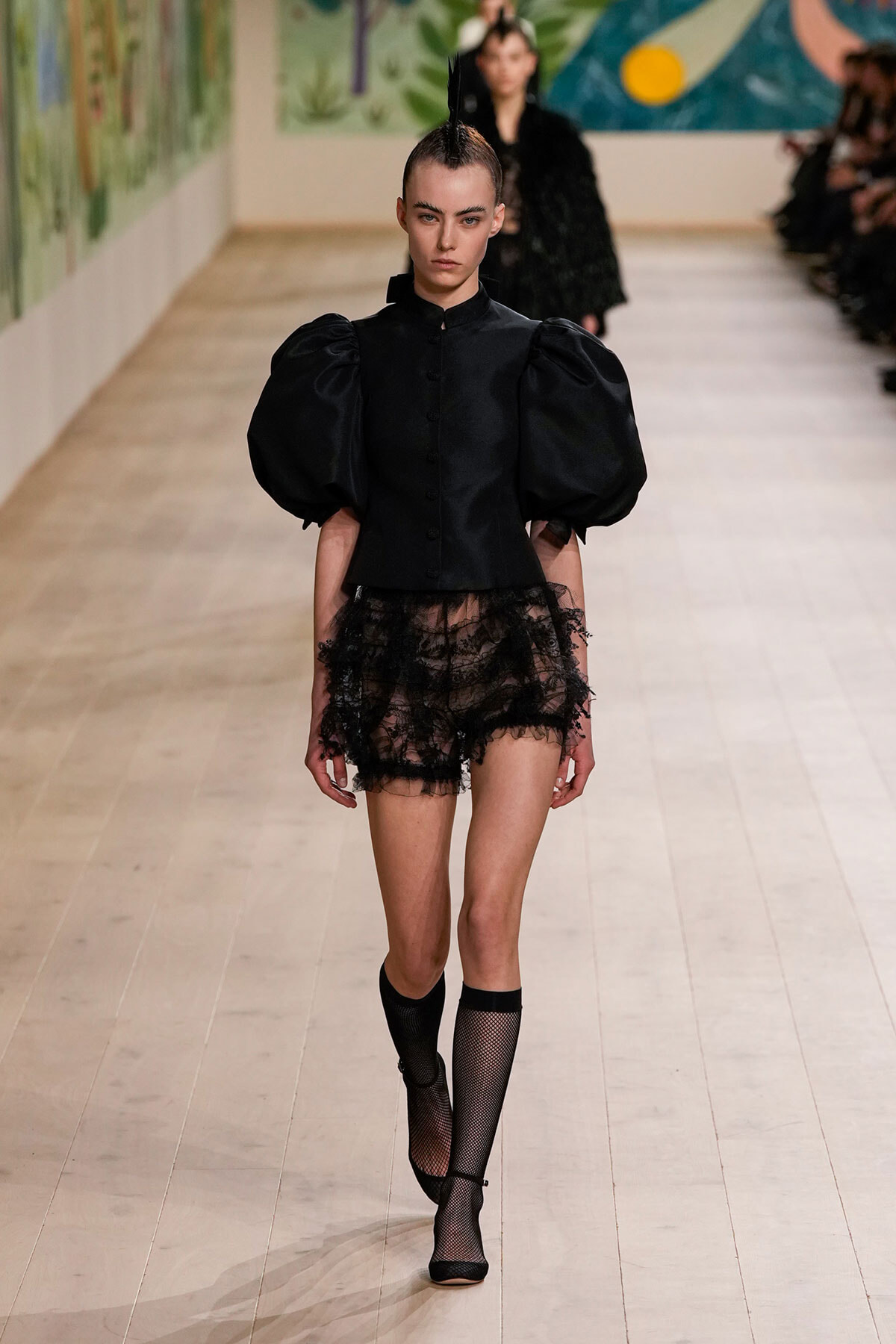 Runway model in a black structured blouse with dramatic puff sleeves, sheer lace shorts, and black fishnet knee-high stockings with heels.