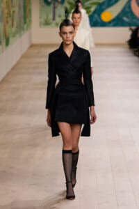 Model walking a runway in a black double-breasted coat-dress with a feather headpiece and fishnet tights, black heels.