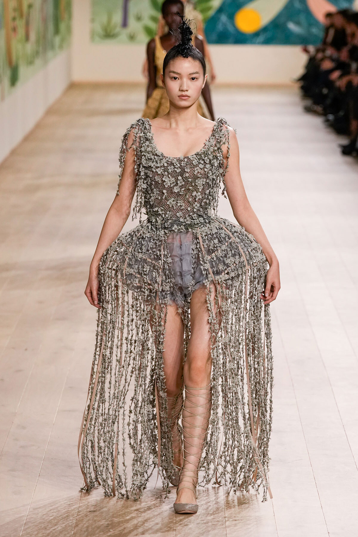 Female model walks runway in a silver-gray floral fringe gown with a sheer bodice and thigh-high lace-up sandals.