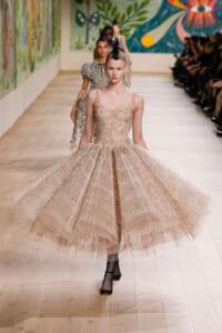 Model walks the runway in a champagne beaded ball gown with a layered tulle skirt and a black feather headpiece on a wooden catwalk.