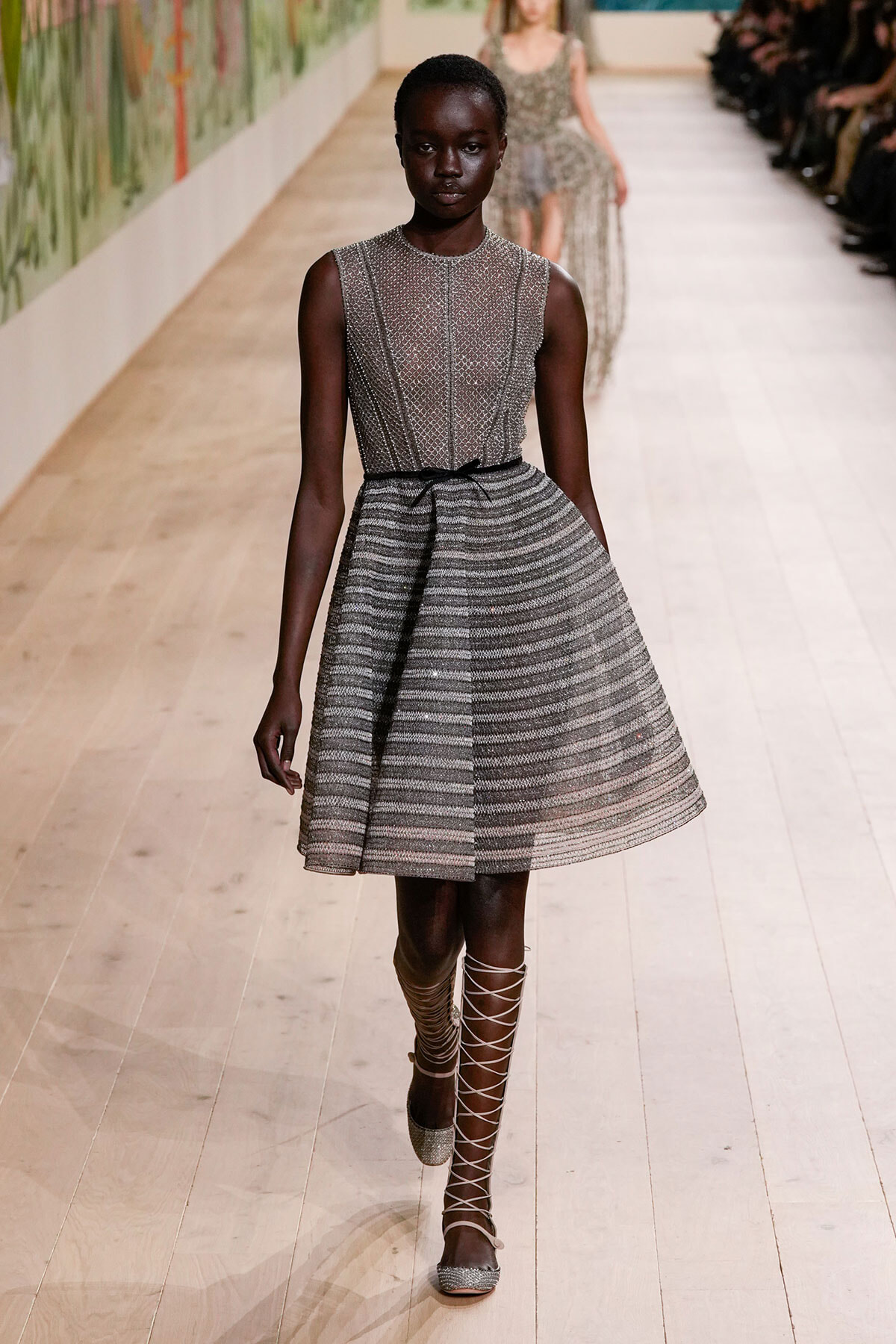Model walking a runway in a silver textured sleeveless dress with a striped A-line skirt and thin belt, pairing silver lace-up sandals.