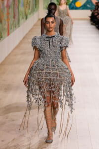 Model walks a runway in a gray, leaf-textured dress with long fringe and strappy lace-up sandals.