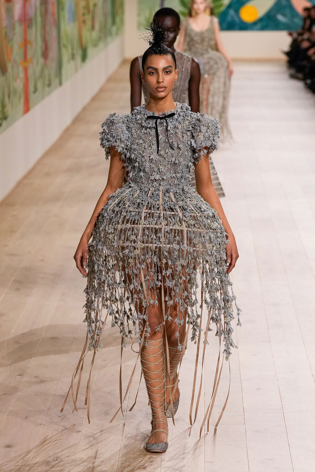 Model walks a runway in a gray, leaf-textured dress with long fringe and strappy lace-up sandals.