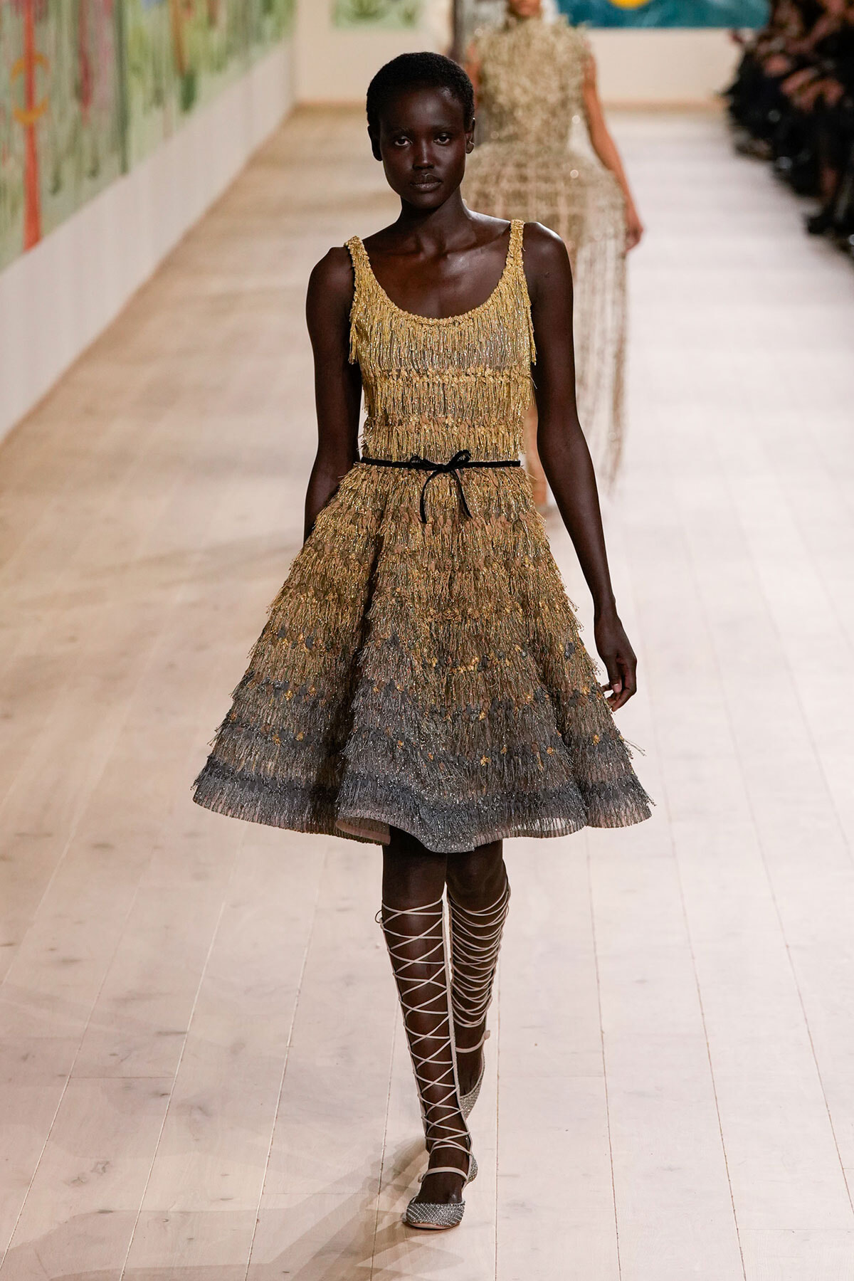 Female model walking the runway in a gold fringe sleeveless midi dress with a thin black belt and lace-up sandals.