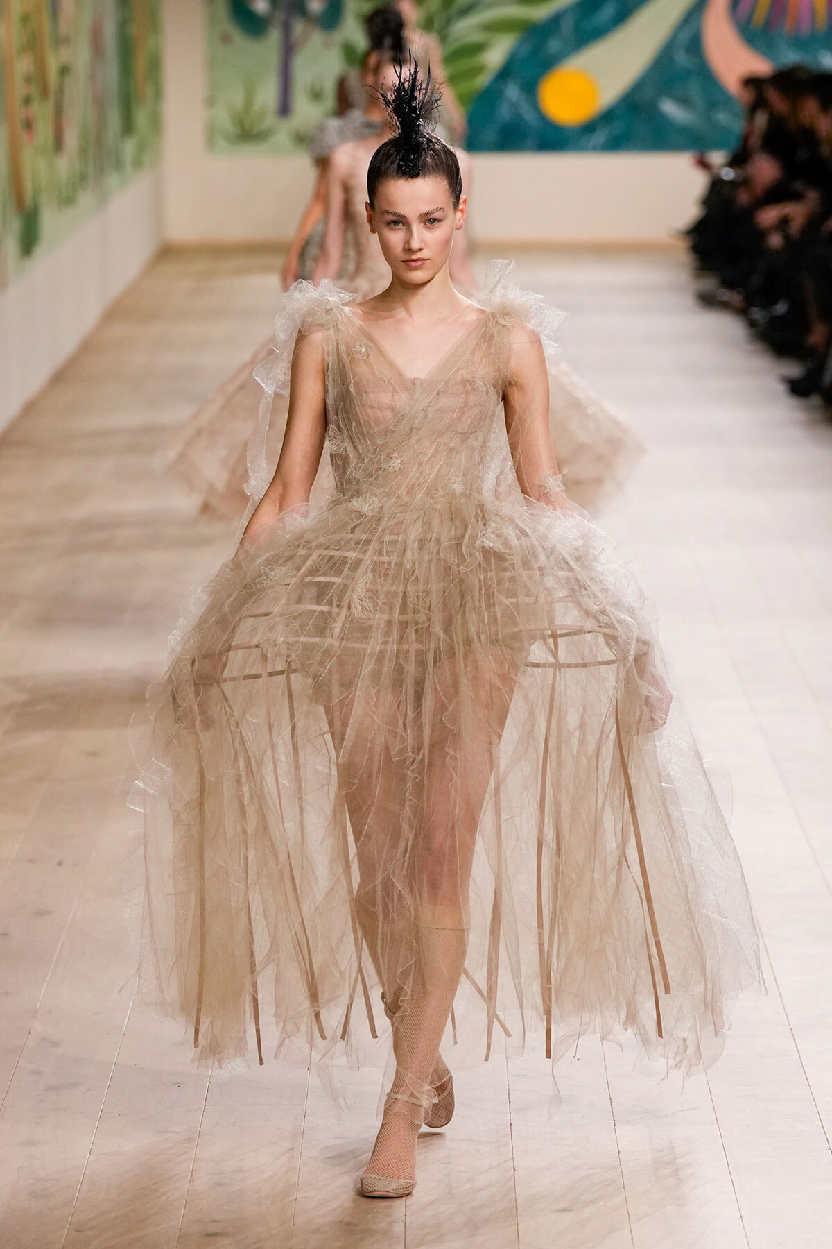 Model walking a runway in a beige sheer tulle gown with layered ruffles and ribbon-like strips, paired with a black feathered headpiece.