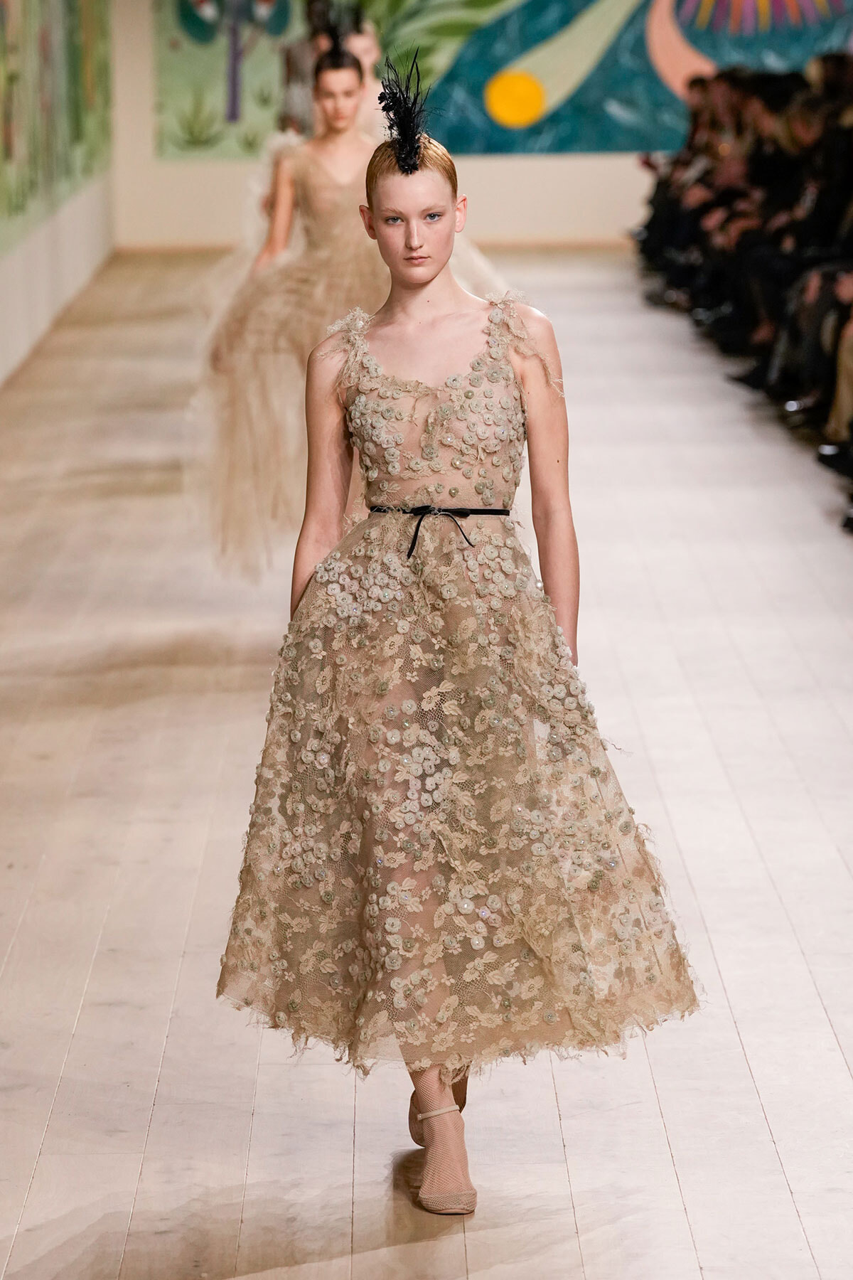Model on a runway in a beige lace ball gown with floral appliqués and a thin black belt. A black feather headpiece tops her hair.