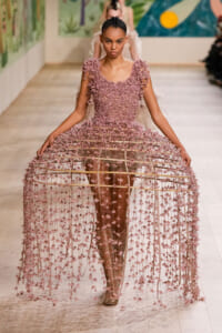 Model walking the runway in a pink petal-covered lattice gown with dangling floral strands forming a voluminous skirt.