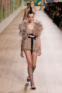 Runway model in a beige, ruffled two-piece outfit with a black sash, feather headpiece, and lace-up sandals.