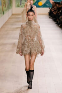 Model on a fashion-show runway wearing a sheer beige lace mini dress with floral embroidery, a ruffled high neck, a feathered headpiece, and black fishnet boots on the catwalk.