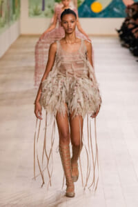 Runway model in a sheer nude top with a feathered fringe skirt and lace-up sandals.