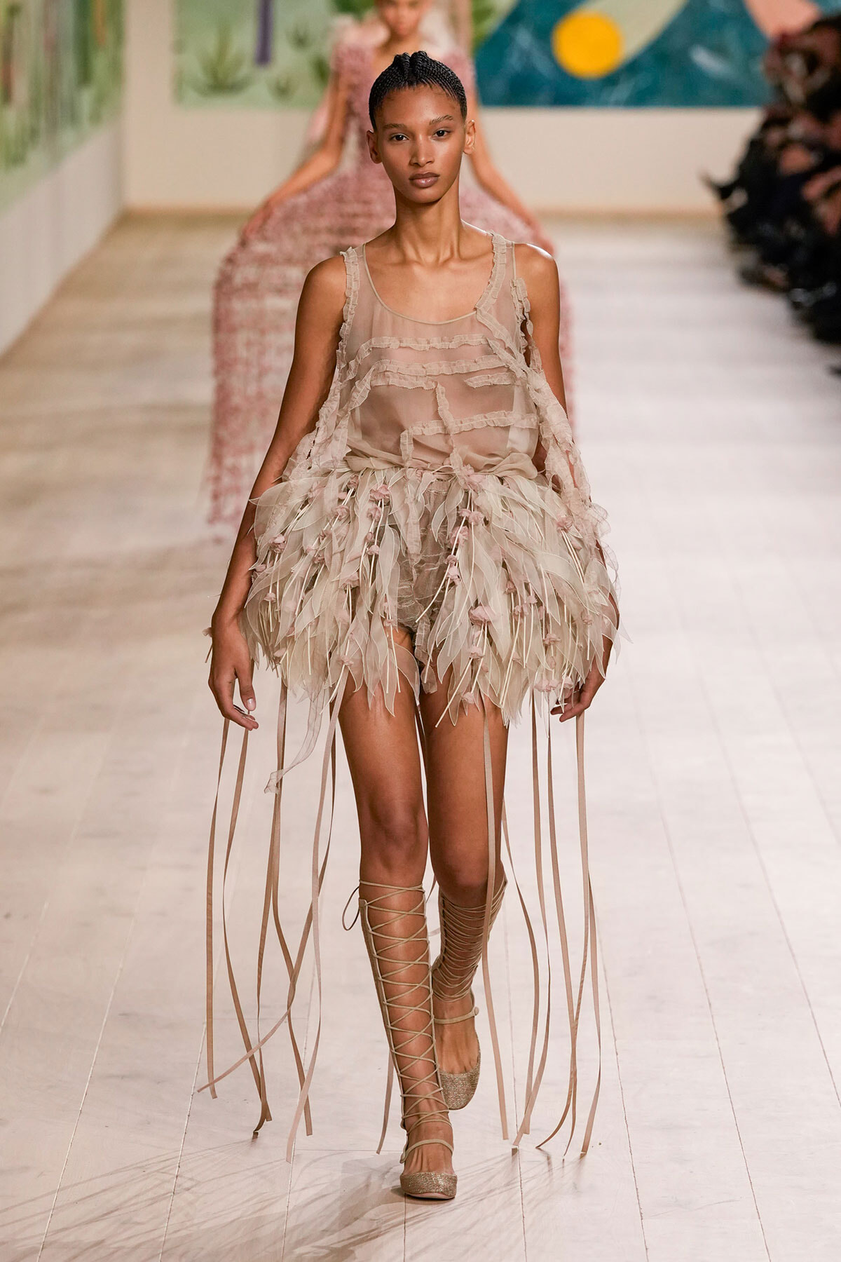 Runway model in a sheer nude top with a feathered fringe skirt and lace-up sandals.