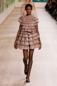 Model walks the runway in a knee-length blush-pink layered ruffle dress with black lace-up gladiator sandals.