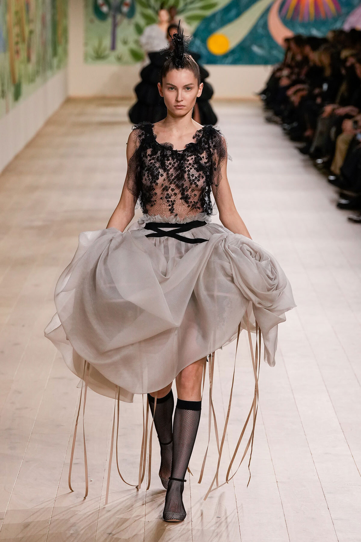 Model walking runway in a sheer black lace top with a voluminous gray skirt and long ribbon details cascading from the hem.