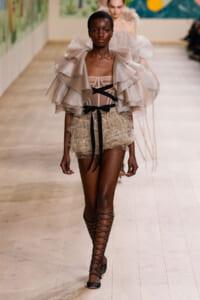 Runway model in a pale pink ruffled cape over a sheer corset and lace shorts, with black ribbon accents and lace-up sandals.