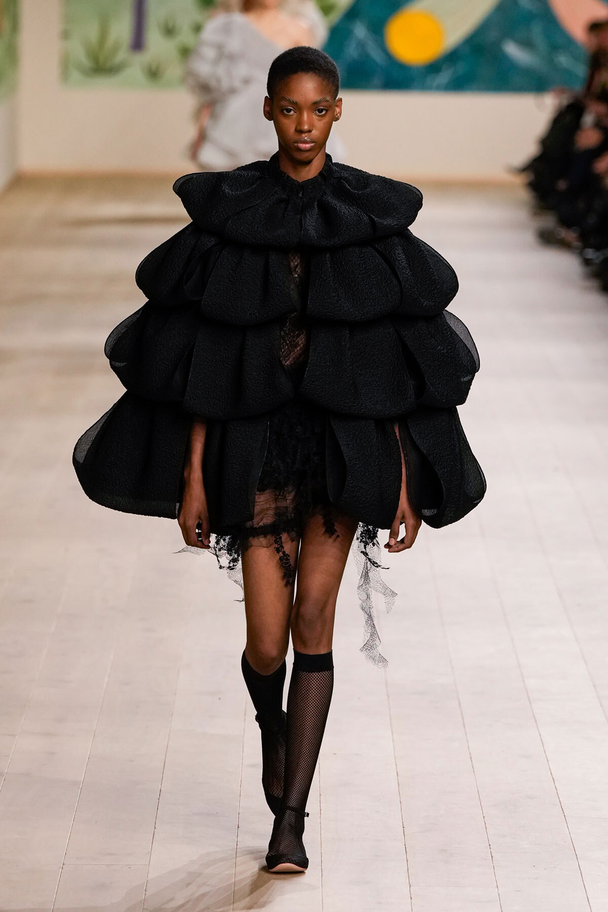 Model on a runway wearing a voluminous black sculptural cape with layered ruffles over a sheer lace dress.