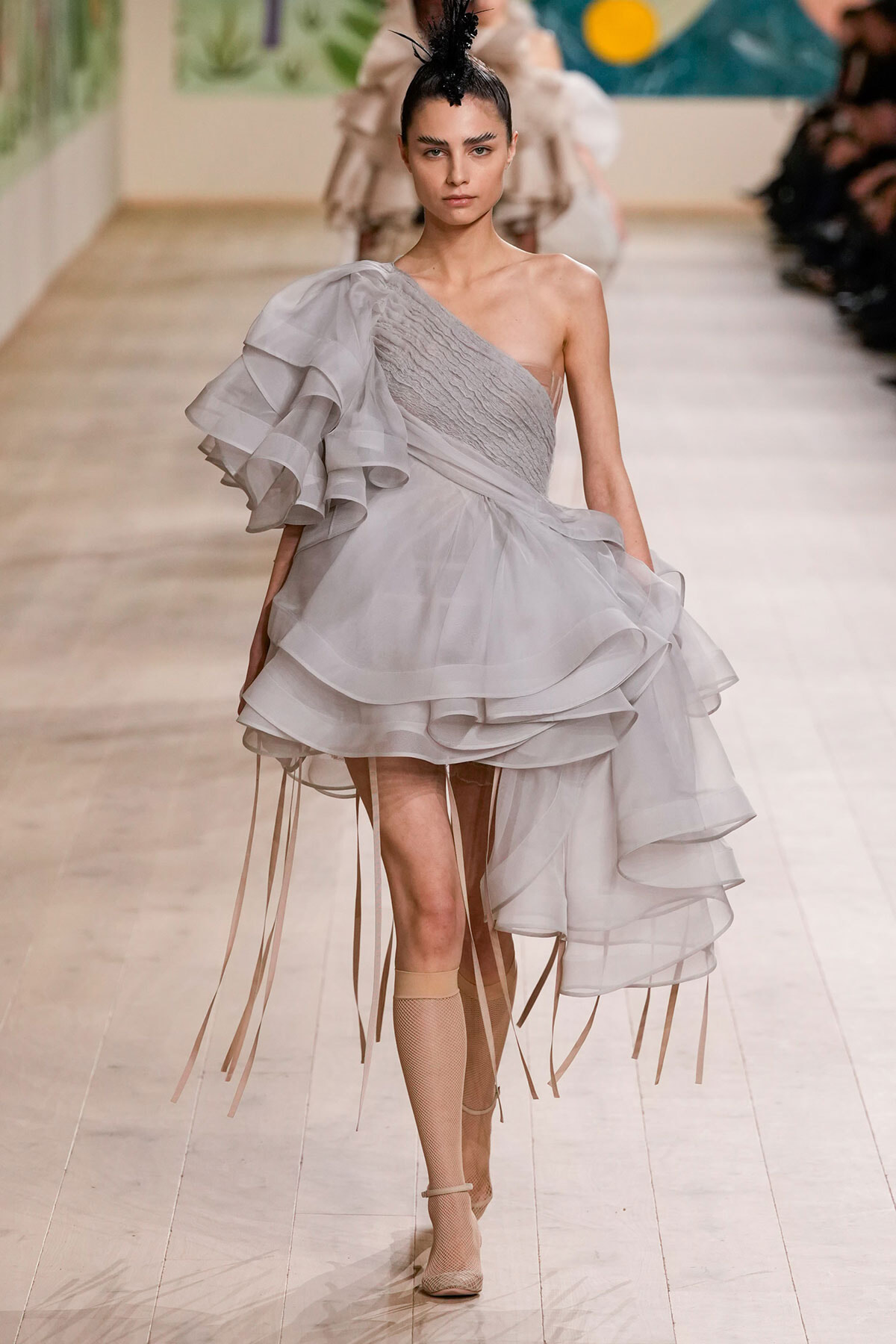 Model walking a runway in a pale gray one-shoulder gown with layered ruffles and fringe details on the skirt.