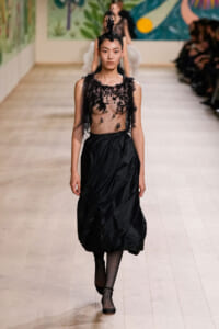 Model walks the runway in a sheer black lace top and a full black skirt with a tall feathered headpiece.