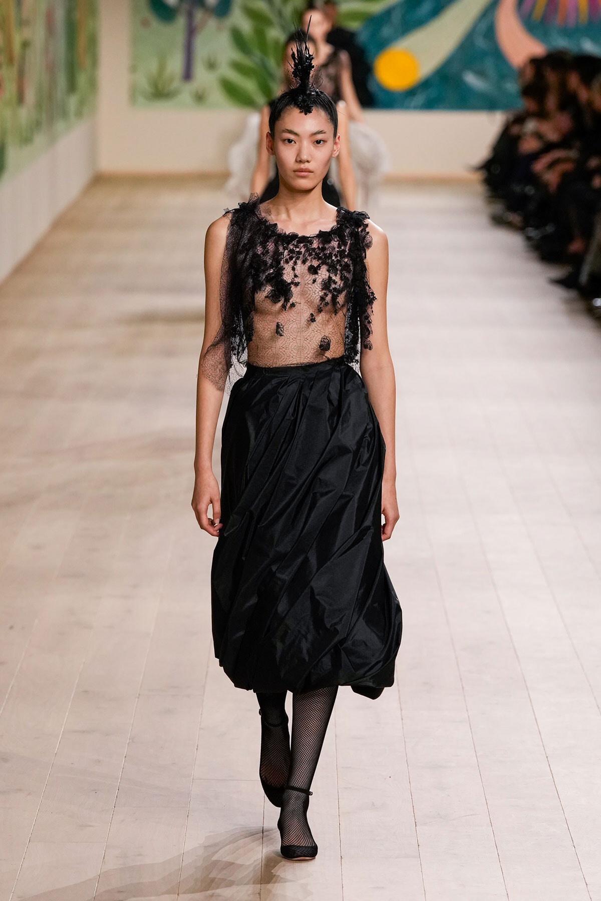 Model walks the runway in a sheer black lace top and a full black skirt with a tall feathered headpiece.