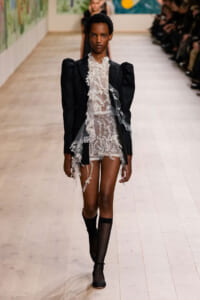 Model walks the runway in a black blazer over a sheer lace dress with knee-high socks.