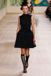 Model on a runway wearing a sleeveless black textured A-line dress with a high neck, paired with black fishnet knee-high socks.