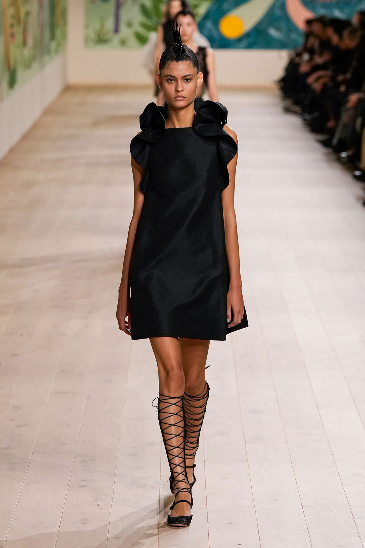 Model walks the runway in a simple black dress with large ruffled shoulders and black lace-up sandals.