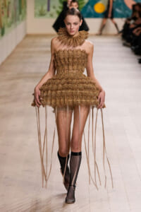 Model on a runway wearing a tan lace dress with a ruffled high neck and long fringe ribbons, plus fishnet socks and flats.