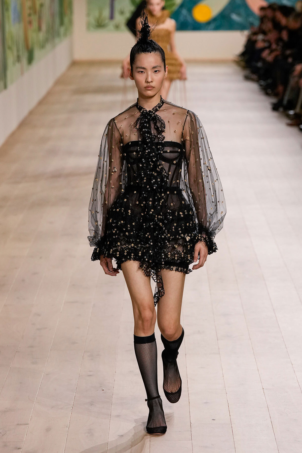 Model walking runway in black embellished sheer dress with star details and high-neck bow, wearing sheer sleeves and fishnet socks.