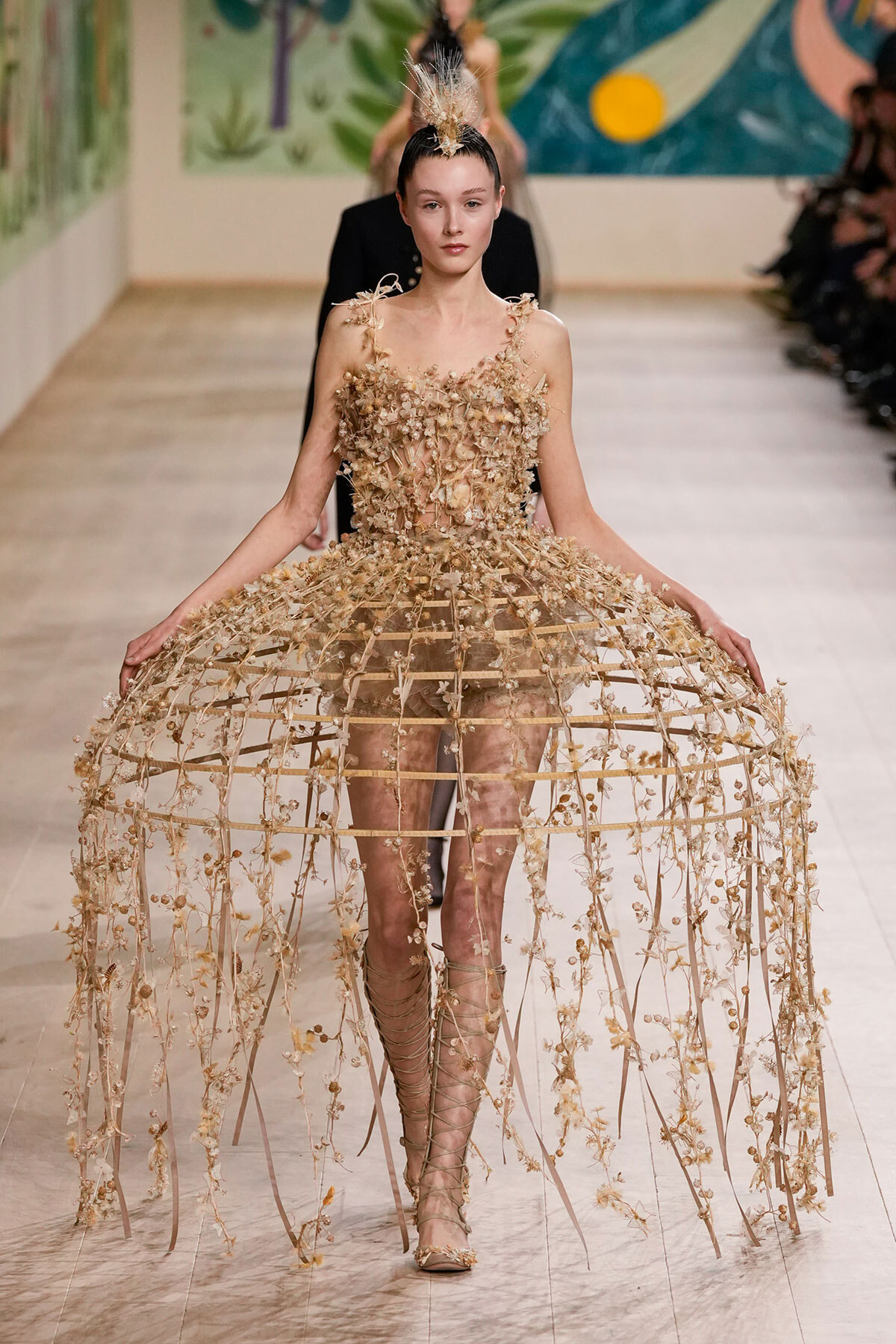 Model walks the runway in a nude-toned lattice cage dress adorned with dried flowers and a tall feathered headpiece.",