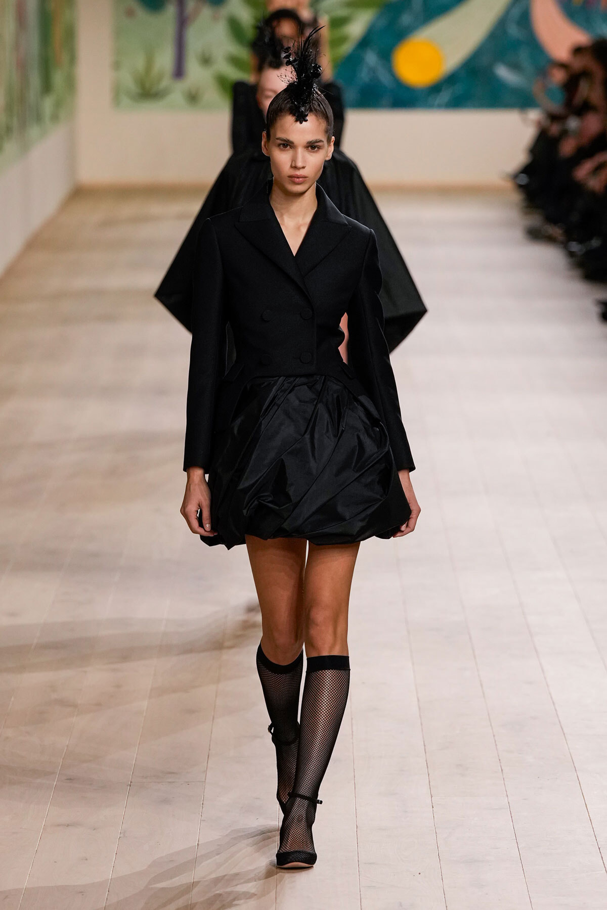 Model walks a fashion show wearing a black double-breasted blazer with a voluminous skirt and feathered headpiece, paired with fishnet knee-high socks.