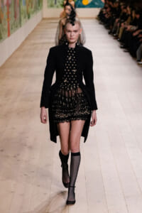 Model walking a runway in a black tailored coat over a sheer floral lace dress with a feathered headpiece and fishnet socks.