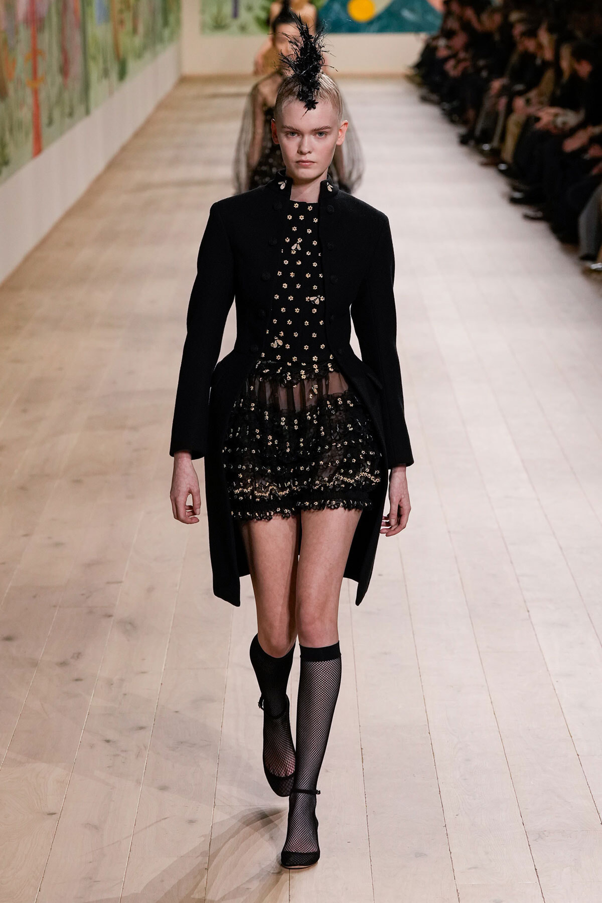Model walking a runway in a black tailored coat over a sheer floral lace dress with a feathered headpiece and fishnet socks.