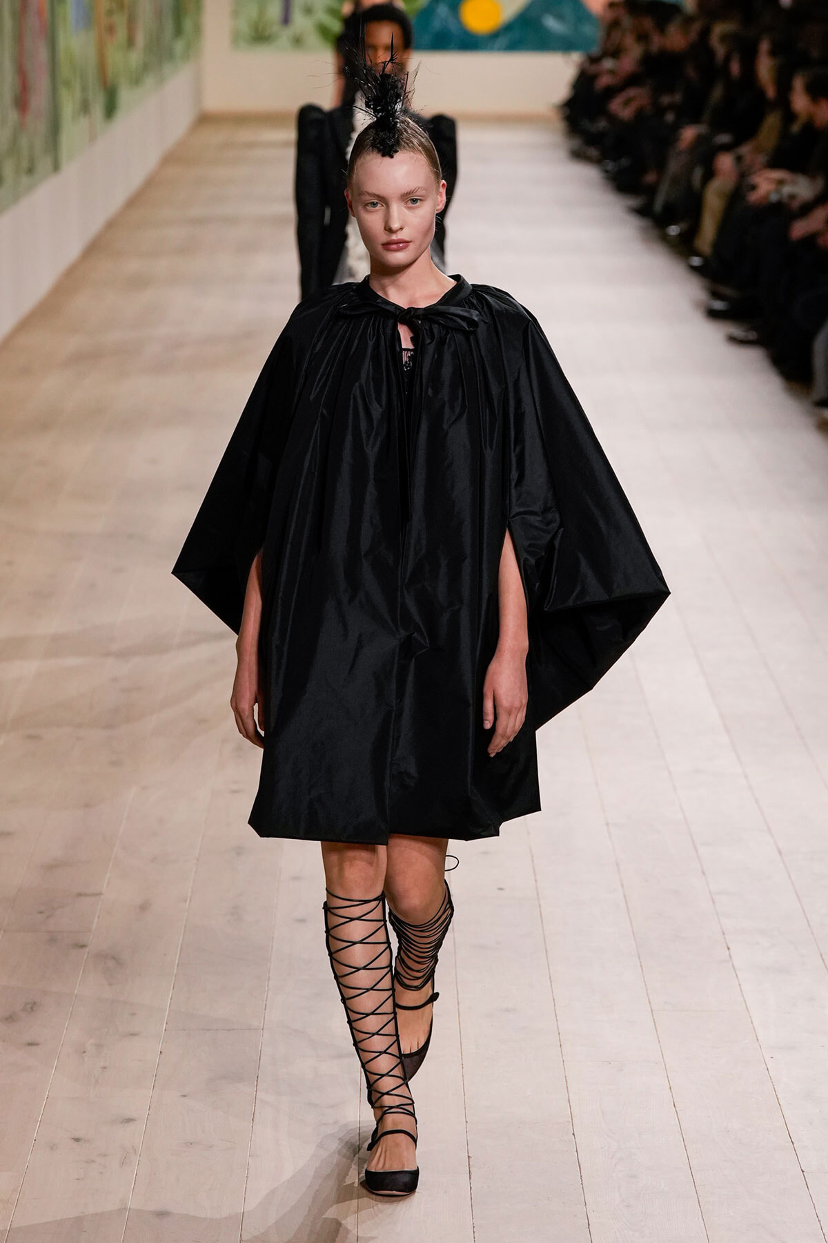 Model walks a fashion show in a loose black cape dress with voluminous sleeves and a feathered headpiece, paired with lace-up sandals.