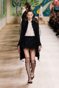 Model on a runway wearing a black feathered-embellished coat over a pale pink buttoned vest, with a black embroidered mini skirt and tall lace-up sandals.
