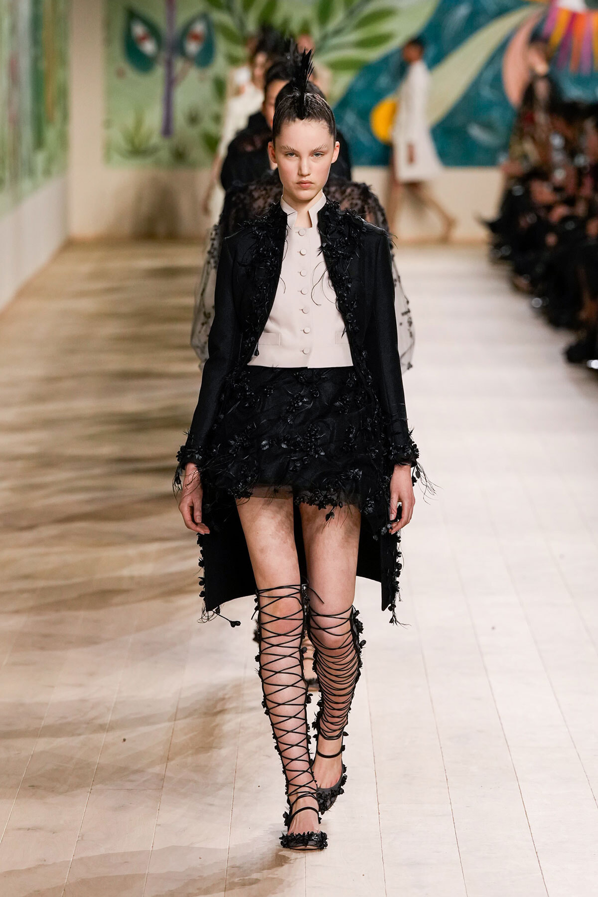 Model on a runway wearing a black feathered-embellished coat over a pale pink buttoned vest, with a black embroidered mini skirt and tall lace-up sandals.