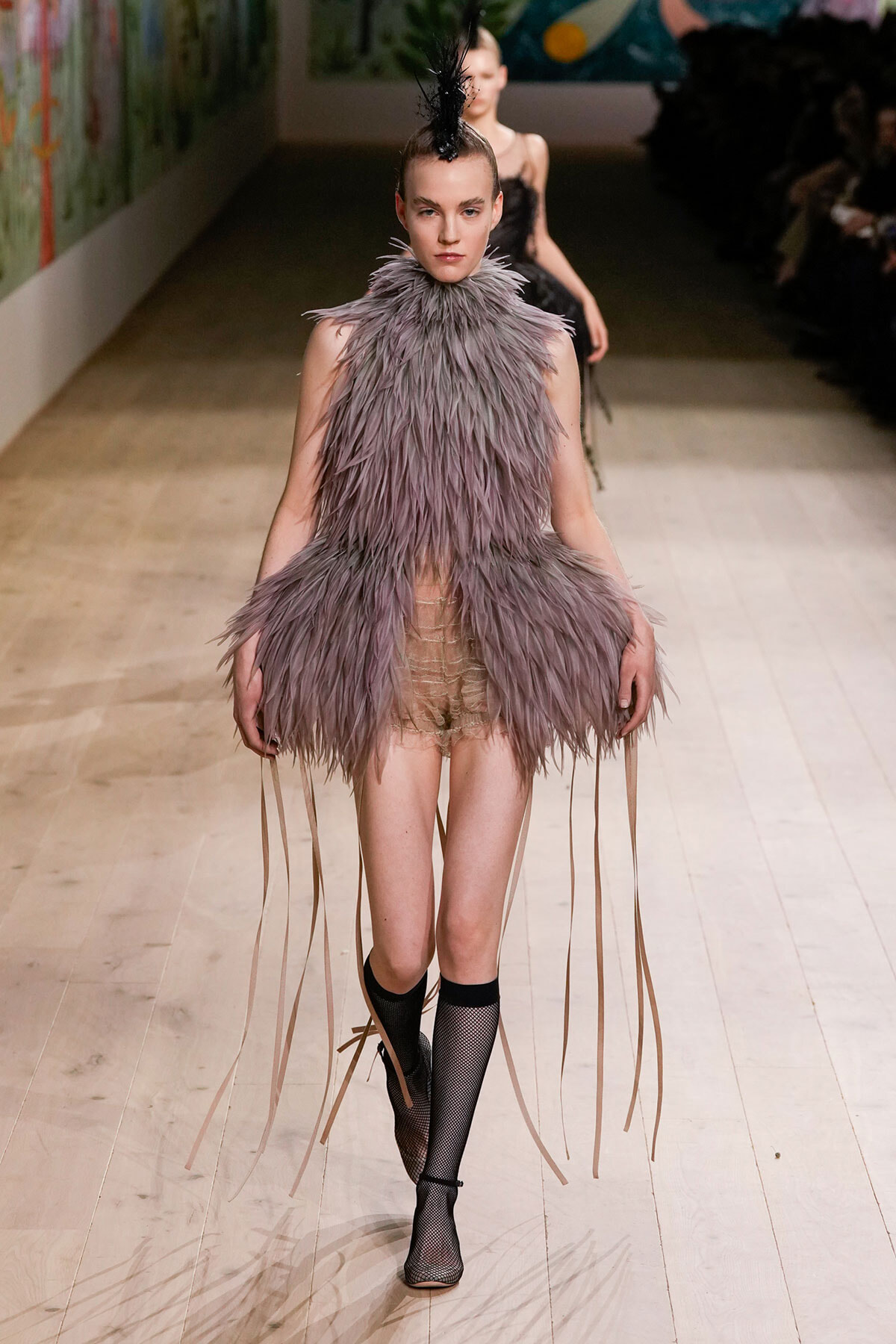 Runway model in a lavender feathered mini dress with a high feathered neck, long fringe strands, and a feather headpiece.