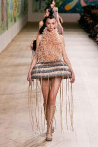 Model walks the runway in a pink feathered halter top and fringe-striped micro skirt with long wrap-around fringe strands, headpiece in black feathers, and lace-up sandals.