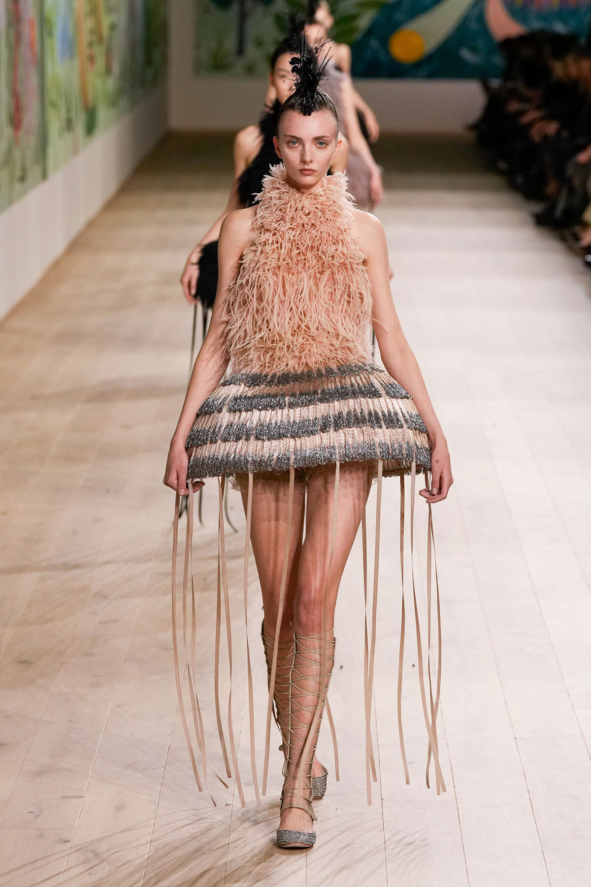 Model walks the runway in a pink feathered halter top and fringe-striped micro skirt with long wrap-around fringe strands, headpiece in black feathers, and lace-up sandals.