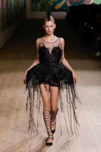 Model walking down a runway in a black lace cocktail dress with sheer bodice and long dangling fringe details, paired with lace-up heels and a feathered headpiece.