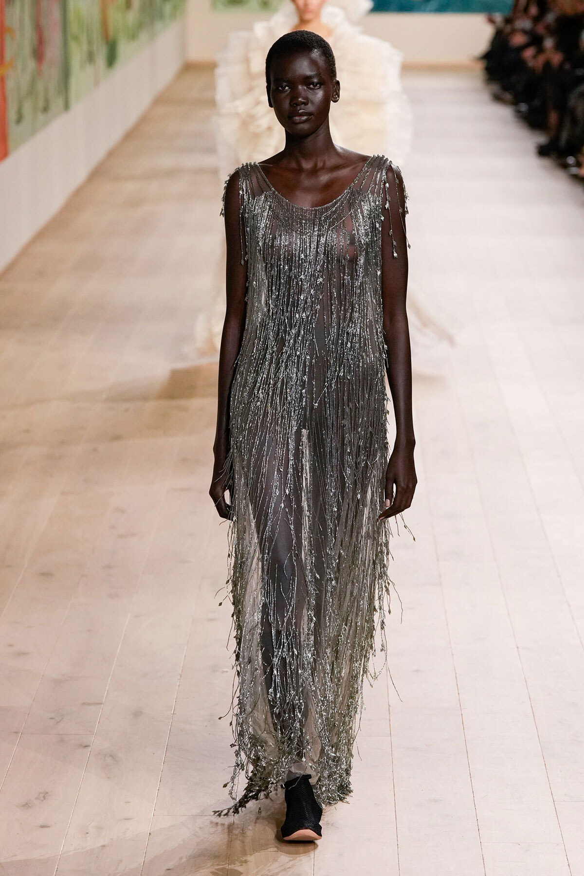 Model walking a runway in a floor-length silver fringe gown with cascading strands and a sheer underlay on a light wooden catwalk.