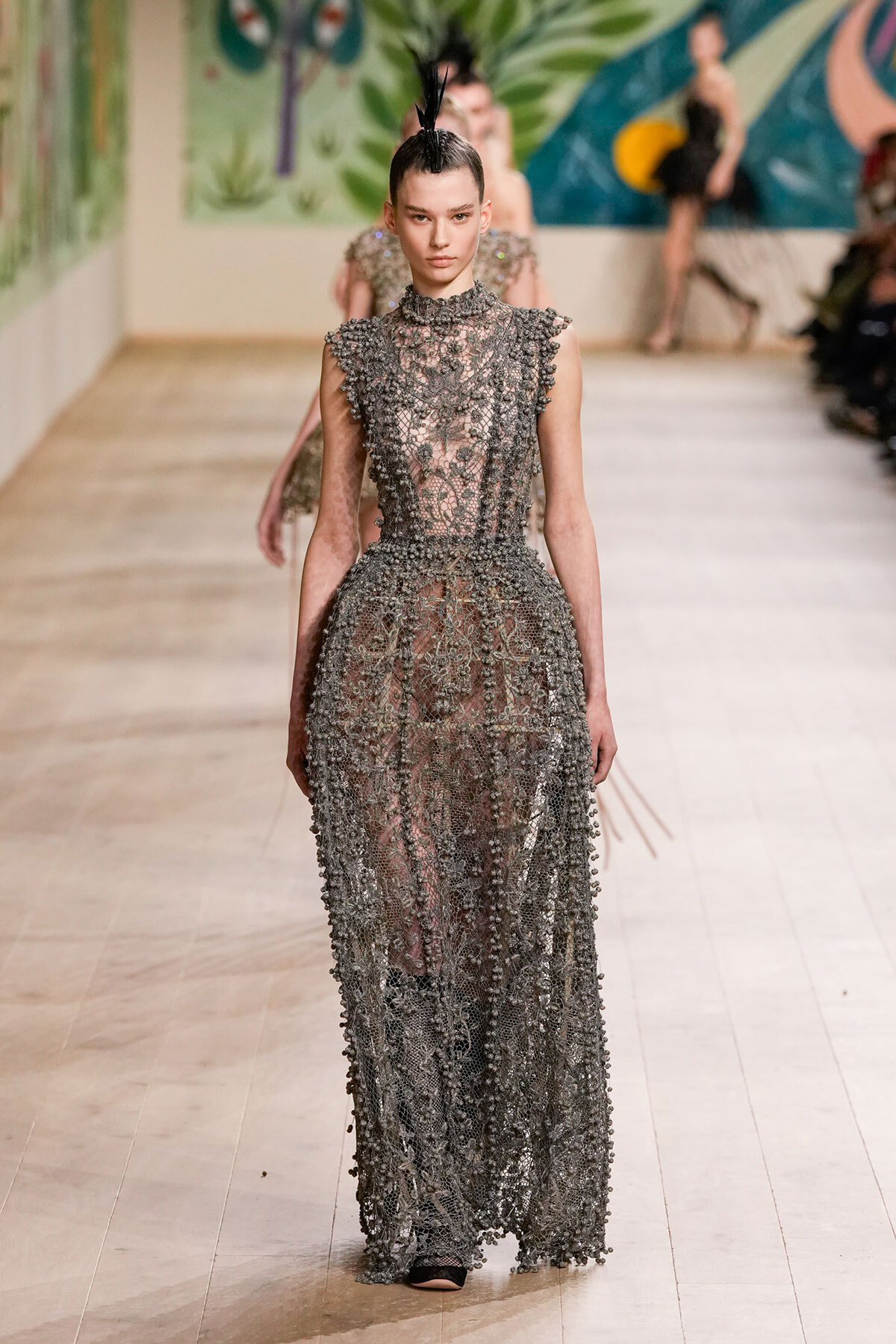 Model walking a runway in a high-neck, floor-length metallic lace gown with intricate beadwork and a spiked updo with a feather accessory.
