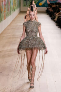 Model walks the runway in a metallic, beaded dress with a full, fringed skirt and lace-up sandals; bold feather headpiece above a slick updo.