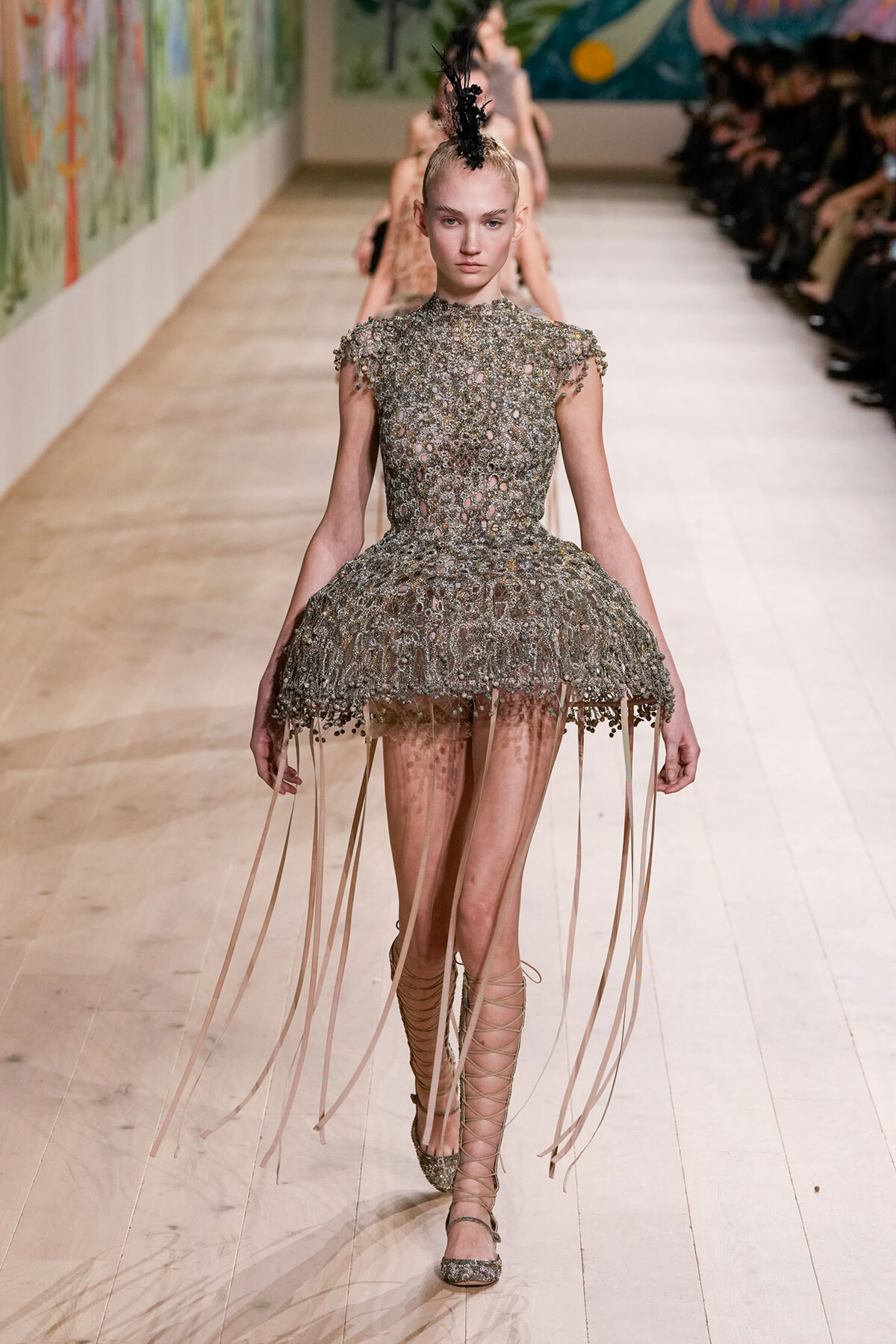 Model walks the runway in a metallic, beaded dress with a full, fringed skirt and lace-up sandals; bold feather headpiece above a slick updo.