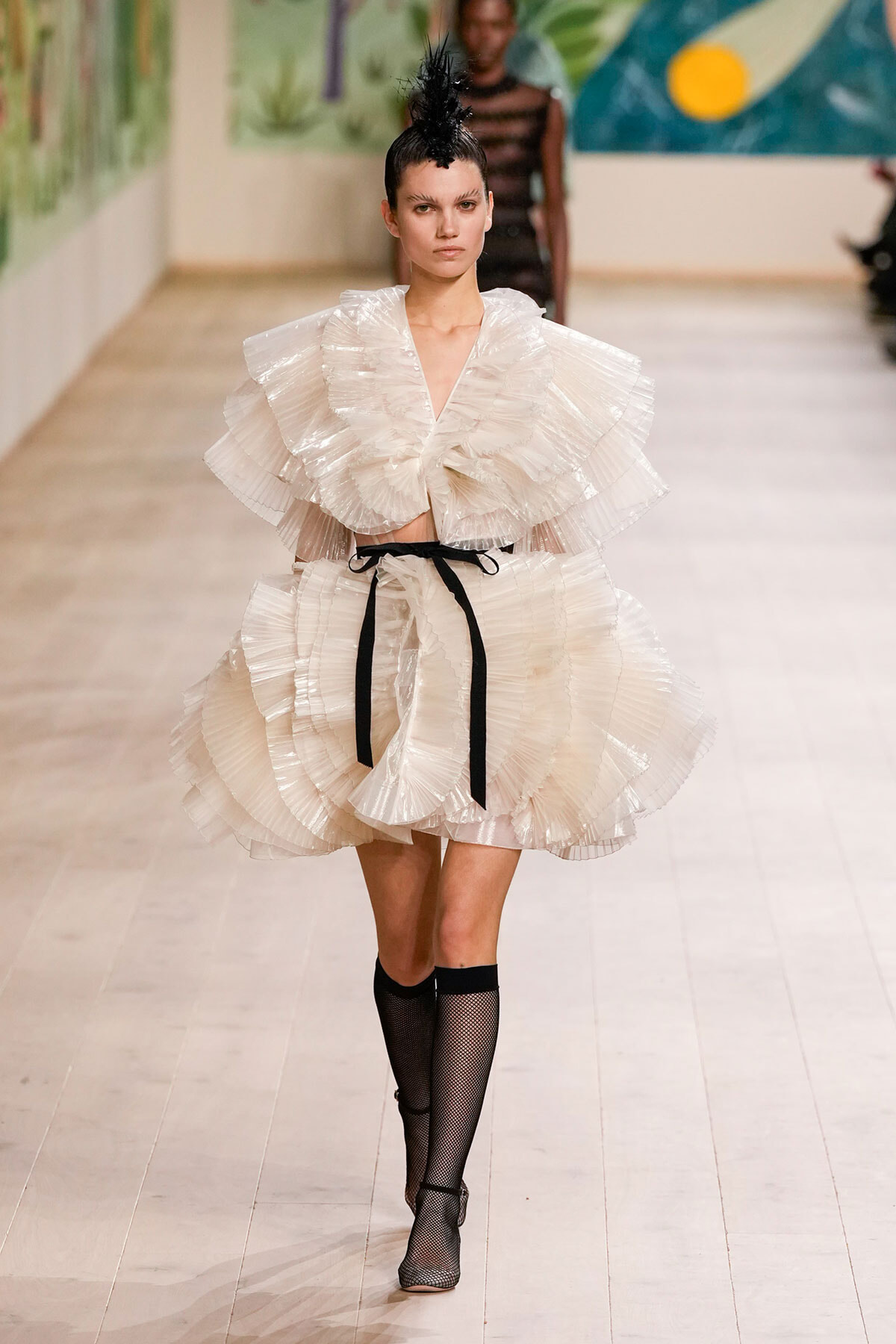 Model walks a runway in a cream, multi-tiered pleated dress with oversized ruffles and a black waist tie.