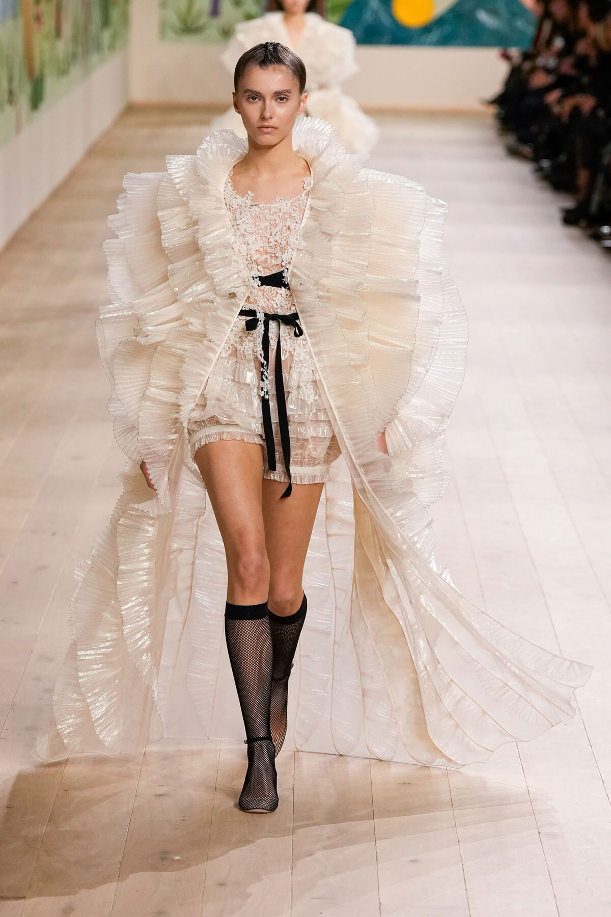 Model walks the runway in a dramatic white pleated cape with ruffles over a lace top and shorts, cinched with a black ribbon belt, and wearing black fishnet knee-high socks.