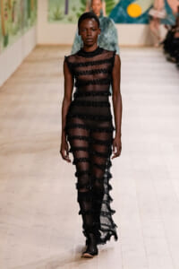 Model walking the runway in a sheer black knit dress with horizontal ruffled bands and high neck, creating a striped, see-through look.