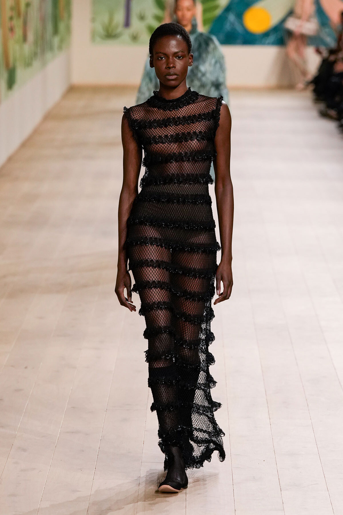 Model walking the runway in a sheer black knit dress with horizontal ruffled bands and high neck, creating a striped, see-through look.