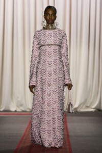 Model on a runway wearing a full-length pink sequin gown with chevron pattern and statement jewelry, carrying a small pink handbag on a red carpet.
