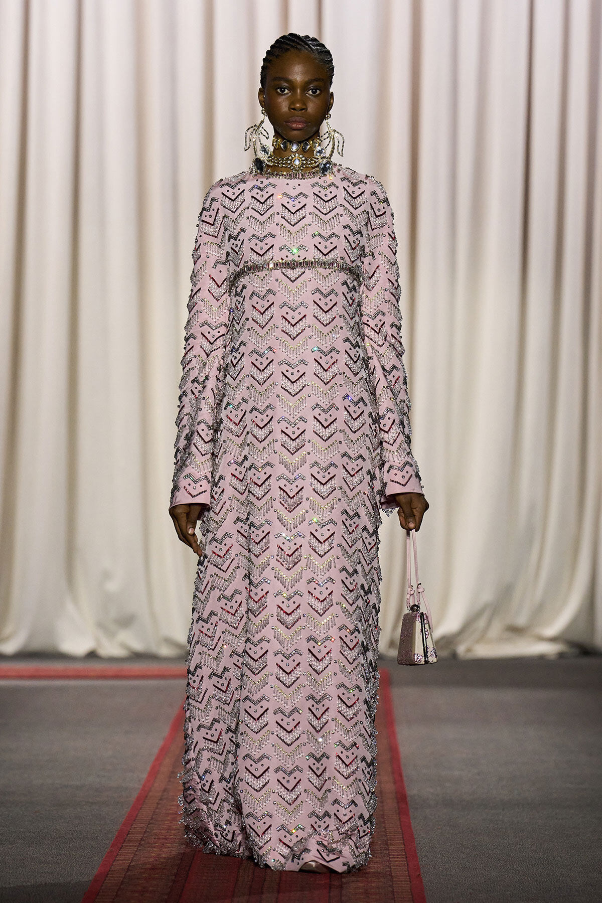 Model on a runway wearing a full-length pink sequin gown with chevron pattern and statement jewelry, carrying a small pink handbag on a red carpet.