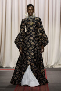 Model on a runway wearing a black and gold floral gown with dramatic puff sleeves and ornate jewelry.