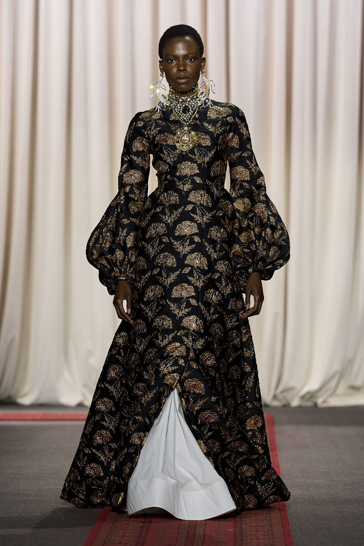 Model on a runway wearing a black and gold floral gown with dramatic puff sleeves and ornate jewelry.